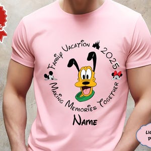 May include: Pale pink t-shirt with a circular design featuring Pluto, Mickey, and Minnie Mouse. The design includes the text "Family Vacation 2025 Making Memories Together" and space for a name. The shirt is a light pink colour.