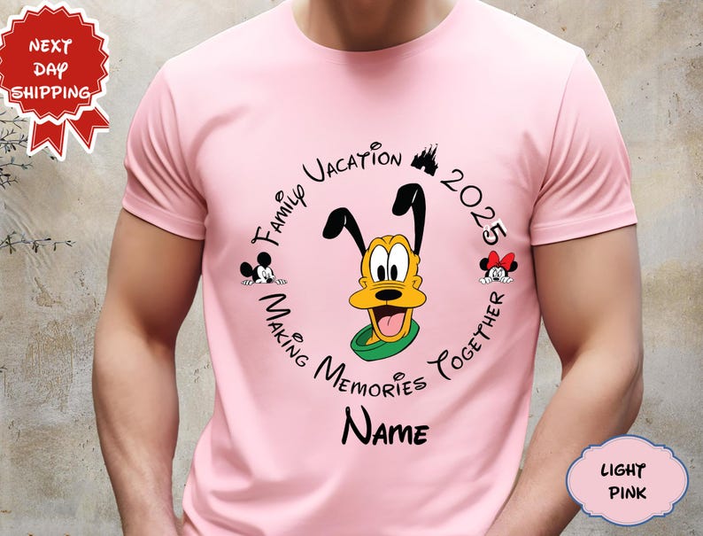 May include: Pale pink t-shirt with a circular design featuring Pluto, Mickey, and Minnie Mouse. The design includes the text "Family Vacation 2025 Making Memories Together" and space for a name. The shirt is a light pink colour.
