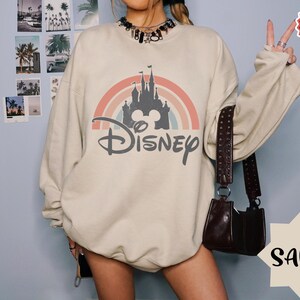 Puede incluir: Una sudadera de cuello redondo color arena con un estampado gr&aacute;fico que muestra la silueta de un castillo gris oscuro bajo un arco&iacute;ris de tonos apagados. La palabra Disney est&aacute; impresa en una fuente cursiva de color gris oscuro.