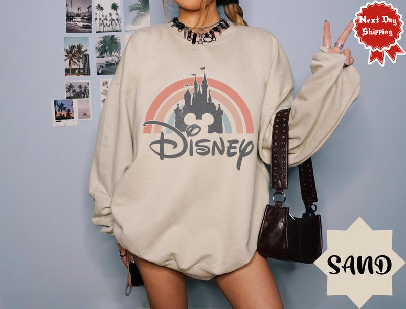 Puede incluir: Una sudadera de cuello redondo color arena con un estampado gr&aacute;fico que muestra la silueta de un castillo gris oscuro bajo un arco&iacute;ris de tonos apagados. La palabra Disney est&aacute; impresa en una fuente cursiva de color gris oscuro.