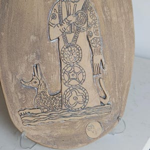 Pode incluir: Placa oval de cerâmica bege, com uma imagem gravada de uma figura com um cocar e barba detalhados. A figura tem padrões intrincados e segura um bastão. Um animal estilizado está aos pés da figura.