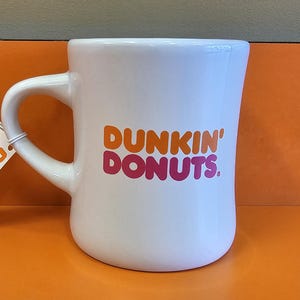 May include: White ceramic mug with the "Dunkin' Donuts" logo in orange and pink. The mug has a curved handle and a tag attached. The mug is on an orange surface.