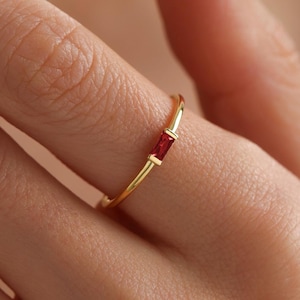 May include: A gold ring with a single red gemstone. The ring is thin and delicate, and the gemstone is a rectangular shape.