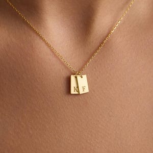 May include: A gold necklace with two small, square pendants, each bearing an engraved initial. The letters "K" and "F" are visible on the pendants. The necklace is worn against a cream-coloured top, offering a simple and elegant aesthetic.