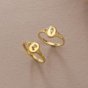 Puede incluir: Dos anillos de sello dorados con diseños florales. Cada anillo presenta una cara ovalada con una flor grabada. Los anillos se exhiben sobre una superficie de tonos neutros, con un fondo suave y borroso.