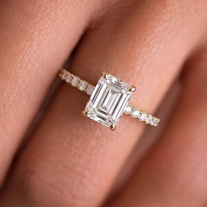 May include: A gold ring featuring a large emerald-cut center stone held by four prongs, with a thin band accented by a row of small round pavé-set diamonds extending along the sides of the shank, creating a bright and reflective appearance.