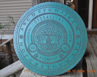 May include: A teal-colored, round Ouija board with engraved letters, numbers, and symbols. The board features the words "YES," "NO," and "REPHRASE." A tree-like design is in the center. The date "09.17.2024" is visible.