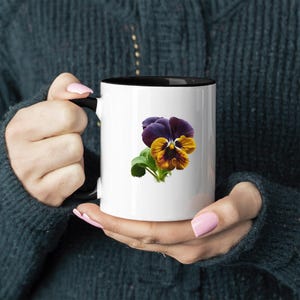 May include: A white ceramic mug with a black rim and handle features a colorful pansy flower illustration. The flower has purple, yellow, and brown petals.