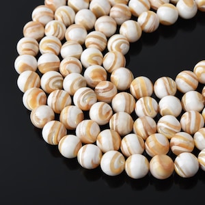 Puede incluir: Una colección de cuentas esféricas dispuestas en una formación curva. Cada cuenta presenta una superficie lisa y pulida con un patrón de remolinos en colores blanco crema y bronceado, característico del material de concha natural con acabado brillante.