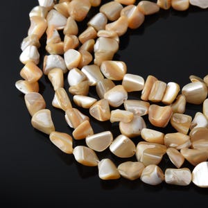May include: A multi-strand necklace composed of irregular, tumbled mother-of-pearl beads in shades of warm beige, cream, and soft peach. The beads feature a smooth, polished finish with a natural iridescent luster, arranged in a curved formation.
