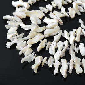 May include: A multi-strand necklace composed of irregular, organic-shaped white mother-of-pearl shell pieces. The shell segments are interspersed with small, clear, faceted crystal beads, creating a textured, layered appearance with a lustrous finish.