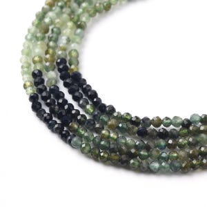 May include: A multi-strand necklace featuring small, faceted, round beads in a gradient of colors. The beads transition from translucent light green and olive tones to deep, opaque black, creating a smooth color shift along the strands.