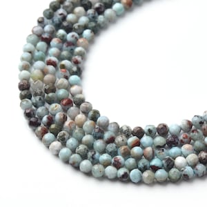 May include: A curved strand of faceted, round beads featuring a mottled pattern of light blue, grey, and dark brown tones. Each individual bead has a polished, multi-faceted surface that reflects light, creating a textured appearance across the strand.