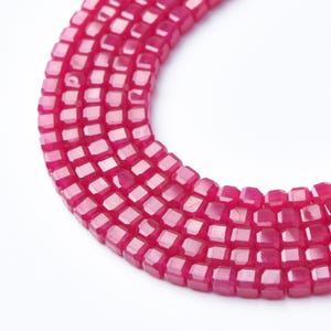 May include: A curved strand of small, square-shaped beads in a vibrant magenta pink color. The beads feature a faceted surface that reflects light, arranged in a dense, multi-row formation that creates a smooth, semi-circular arc shape.