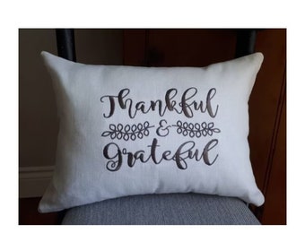 May include: White throw pillow with the text "Thankful & Grateful" embroidered in brown cursive font. The text is separated by a small line of brown embroidered leaves.