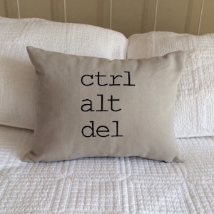 Peut inclure: Un coussin beige avec le texte "ctrl alt del" brodé en noir.
