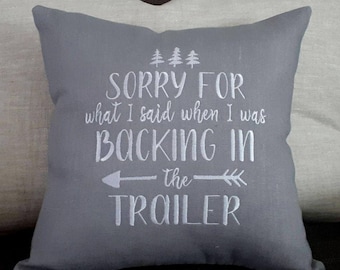 May include: Gray throw pillow with white embroidered text that reads "Sorry for what I said when I was backing in the trailer". The text is accompanied by a small tree graphic and an arrow pointing to the right.