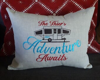 May include: A gray linen pillow with embroidered text that reads "The Thiers Adventure Awaits" and a black and white illustration of a pop-up camper with the year 2020 printed on the side. The text "Adventure" is embroidered in blue and the text "Awaits" is embroidered in red.