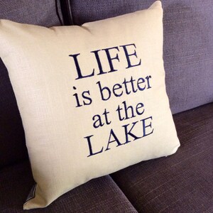 May include: A beige throw pillow with the text "LIFE is better at the LAKE" embroidered in navy blue.