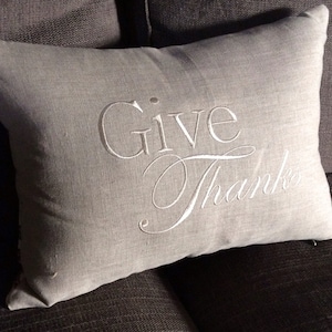 May include: A gray linen throw pillow with white embroidery that reads "Give Thanks".