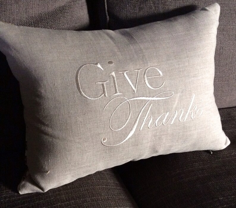 May include: A gray linen throw pillow with white embroidery that reads "Give Thanks".
