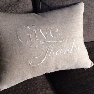 May include: A gray linen throw pillow with white embroidery that reads "Give Thanks".