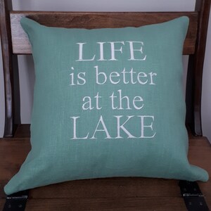 May include: A teal green throw pillow with white embroidered text that reads "LIFE is better at the LAKE".
