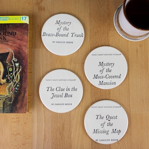 May include: Four white coasters with black text featuring the titles of Nancy Drew mystery books. The titles are "Mystery of the Brass-Bound Trunk", "The Clue in the Jewel Box", "Mystery of the Moss-Covered Mansion", and "The Quest of the Missing Map".