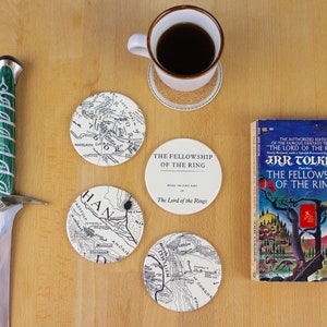 May include: Four white ceramic coasters with black and white maps of Middle-earth from The Lord of the Rings. The coasters feature the text "The Fellowship of the Ring" and "The Land of the Rings". A book titled "The Fellowship of the Ring" by J.R.R. Tolkien is also pictured.