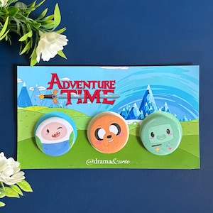 Puede incluir: Juego de tres botones de personajes de Adventure Time. Cada botón presenta un personaje diferente: Finn, Jake y BMO. Los botones están en una tarjeta con el logotipo de Adventure Time y un fondo de paisaje. La tarjeta tiene el texto "@drama&carte".