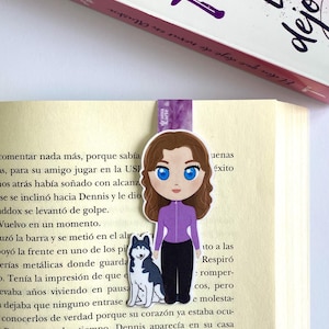 Puede incluir: Un marcapáginas morado y blanco con una ilustración de dibujos animados de una mujer con cabello castaño y ojos azules que lleva una camisa morada y pantalones negros. Está de pie junto a un perro blanco y negro. El marcapáginas está colocado en un libro con el texto "Ali Keller" y "El día que dejé de querer" visible.