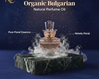 May include: A perfume bottle with a floral design sits on a marble block, with the text "Organic Bulgarian Natural Perfume Oil." The bottle is filled with a light brown liquid and has a clear, flower-shaped stopper. The background is a dark blue with white specks.