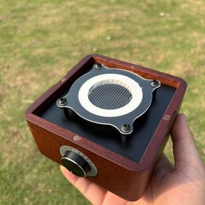 Peut inclure: Un appareil audio carré en bois avec un dessus noir et une grille de haut-parleur circulaire. L'appareil a un gros bouton sur le côté et est tenu dans une main sur un fond vert.