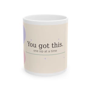 Könnte beinhalten: Weiße Keramik-Tasse mit dem Text "You got this." in Schwarz und "one sip at a time" in kleinerer Schrift. Die Tasse hat einen beigefarbenen Hintergrund mit lila und rosa Akzenten.