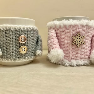 May include: Two handmade crochet mug cozies. One is gray with two gold buttons, resembling a sweater. The other is pink with white trim and a wooden snowflake accent. These cozy accessories are designed to fit around a standard mug.