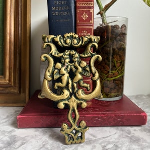 May include: A gold-toned metal bookend with an ornate design featuring two cherubs holding a heart. The bookend is sitting on top of a red book with the title "The Republic" visible.
