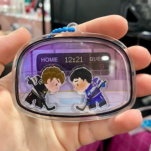 May include: A clear, rectangular keychain with a hockey theme. The keychain features two cartoon hockey players facing each other on an ice rink. The scoreboard reads "HOME 12:21 GUES 9". The keychain has a blue beaded chain.