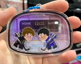Könnte beinhalten: Ein klarer, rechteckiger Schlüsselanhänger mit Hockey-Motiv. Der Schlüsselanhänger zeigt zwei Cartoon-Hockeyspieler, die sich auf einer Eisbahn gegenüberstehen. Die Anzeigetafel zeigt "HOME 12:21 GUES 9". Der Schlüsselanhänger hat eine blaue Perlenkette.