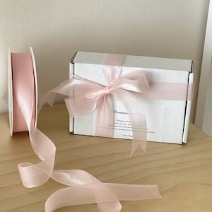 Könnte beinhalten: Eine weiße Geschenkbox mit einem rosa Band und einem rosa durchsichtigen Band. Die Box hat ein Etikett mit der Aufschrift "Romance Gift Box" und enthält eine Botschaft über das größte Glück, zu lieben und geliebt zu werden.