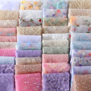Könnte beinhalten: Eine Sammlung verschiedener Stoffmuster, die eine Reihe von Farben und Texturen zeigen. Die Stoffe umfassen Spitze, Tüll und gestickte Designs mit floralen und Pailletten-Verzierungen. Farben sind Weiß, Rosa, Lila und Beige.