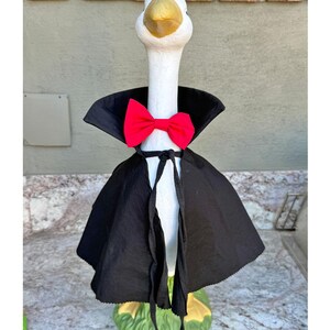 May include: A white goose figurine wearing a black cape with a high collar and a bright red bow tie. The goose has a yellow beak and is standing on a green base. The cape is secured at the neck with black fabric ties that hang down the front.