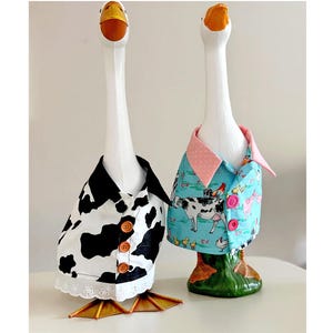 May include: Two white goose figurines with orange beaks. The left goose wears a black and white cow-print shirt with three brown buttons and a white eyelet lace trim. The right goose wears a light blue shirt with farm animal prints and pink collar.