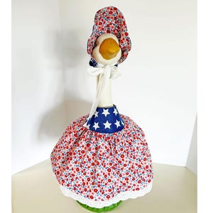 May include: A decorative goose figurine wearing a patriotic outfit. The outfit includes a red floral print dress with a blue star-studded collar and matching bonnet. The goose has a white neck and a yellow beak.