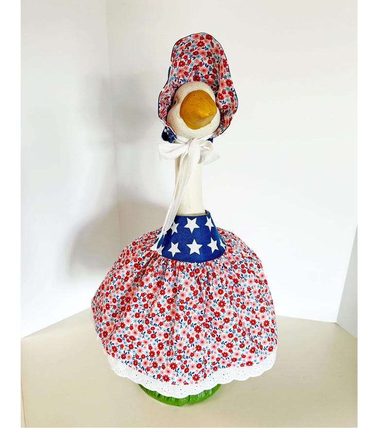 May include: A decorative goose figurine wearing a patriotic outfit. The outfit includes a red floral print dress with a blue star-studded collar and matching bonnet. The goose has a white neck and a yellow beak.