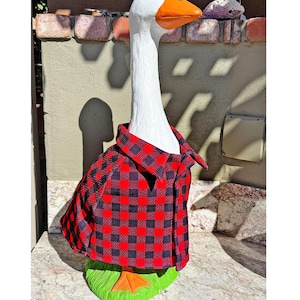 May include: A white goose figurine with an orange beak, wearing a red and black buffalo plaid shirt. The goose stands on a green base. The shirt features a classic collar and a button-down front design, covering the torso of the bird statue.