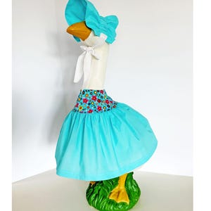 May include: A decorative goose statue wearing a turquoise bonnet and skirt. The skirt has a floral pattern at the waist. The goose has a white neck, a yellow beak, and is standing on a green base.