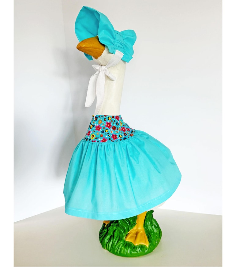 May include: A decorative goose statue wearing a turquoise bonnet and skirt. The skirt has a floral pattern at the waist. The goose has a white neck, a yellow beak, and is standing on a green base.