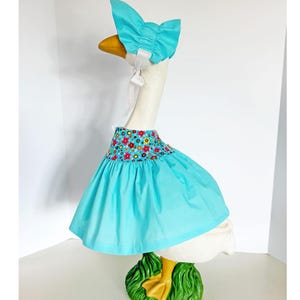 May include: A decorative goose figurine wearing a turquoise dress and bonnet. The dress features a floral patterned bodice. The goose stands on a green base with yellow feet. The bonnet is tied with white ribbons.