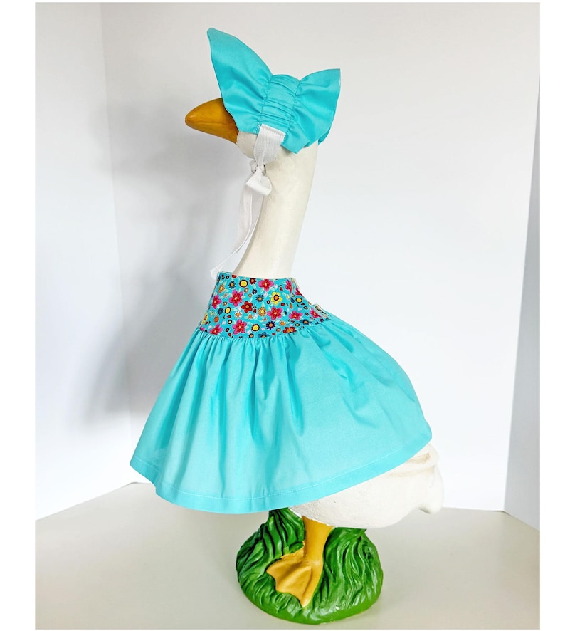 May include: A decorative goose figurine wearing a turquoise dress and bonnet. The dress features a floral patterned bodice. The goose stands on a green base with yellow feet. The bonnet is tied with white ribbons.