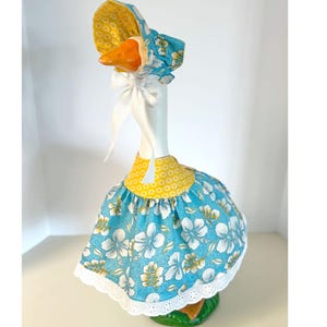 May include: A decorative goose figurine wearing a blue floral print dress and bonnet with yellow accents. The dress has a white lace trim. The goose has an orange beak and is standing on a green base.
